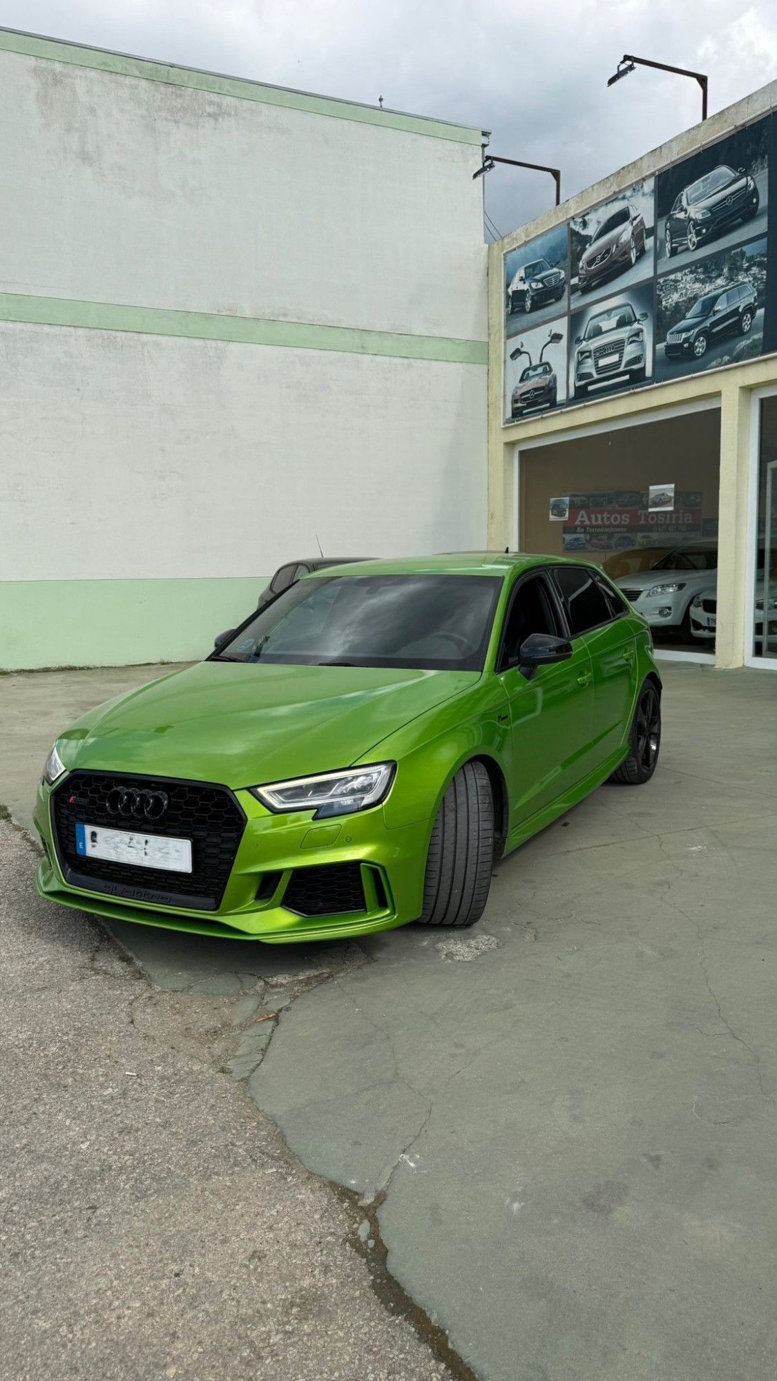 Un sedán deportivo de color verde brillante estacionado frente a un garaje con letreros de autos en la pared.