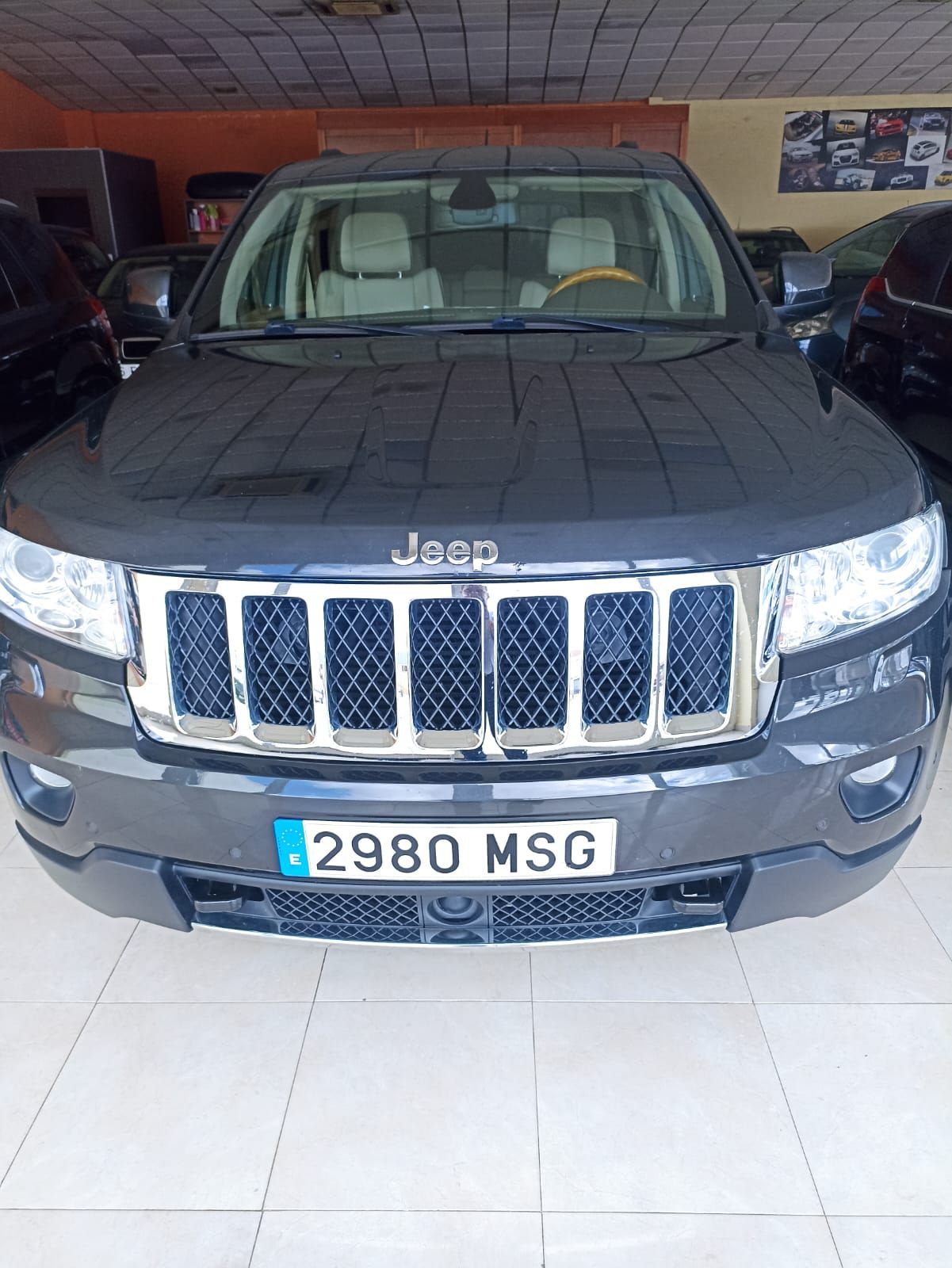 Jeep SUV gris oscuro estacionado en el interior de una sala de exposición, vista frontal con parrilla cromada 