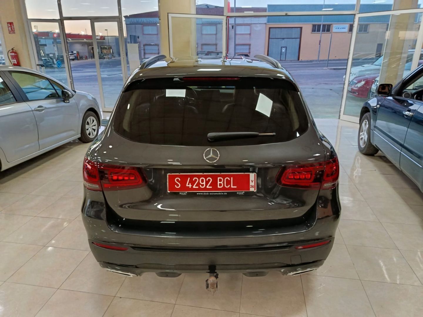 Vista trasera de un SUV oscuro con matrícula roja estacionado en una sala de exposición entre dos coches.