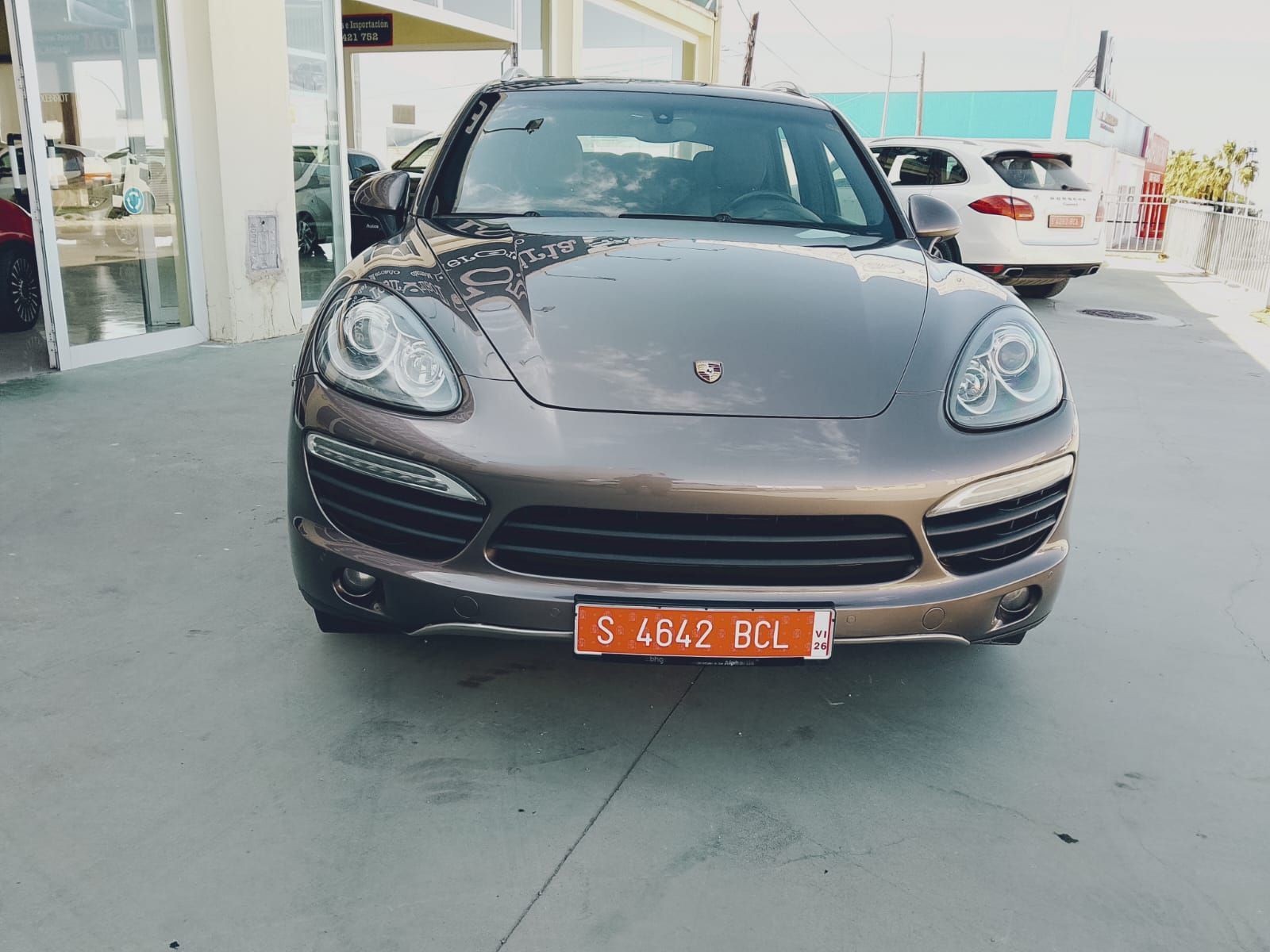 Coche deportivo Porsche gris aparcado frente a un concesionario, vista frontal con matrícula naranja.