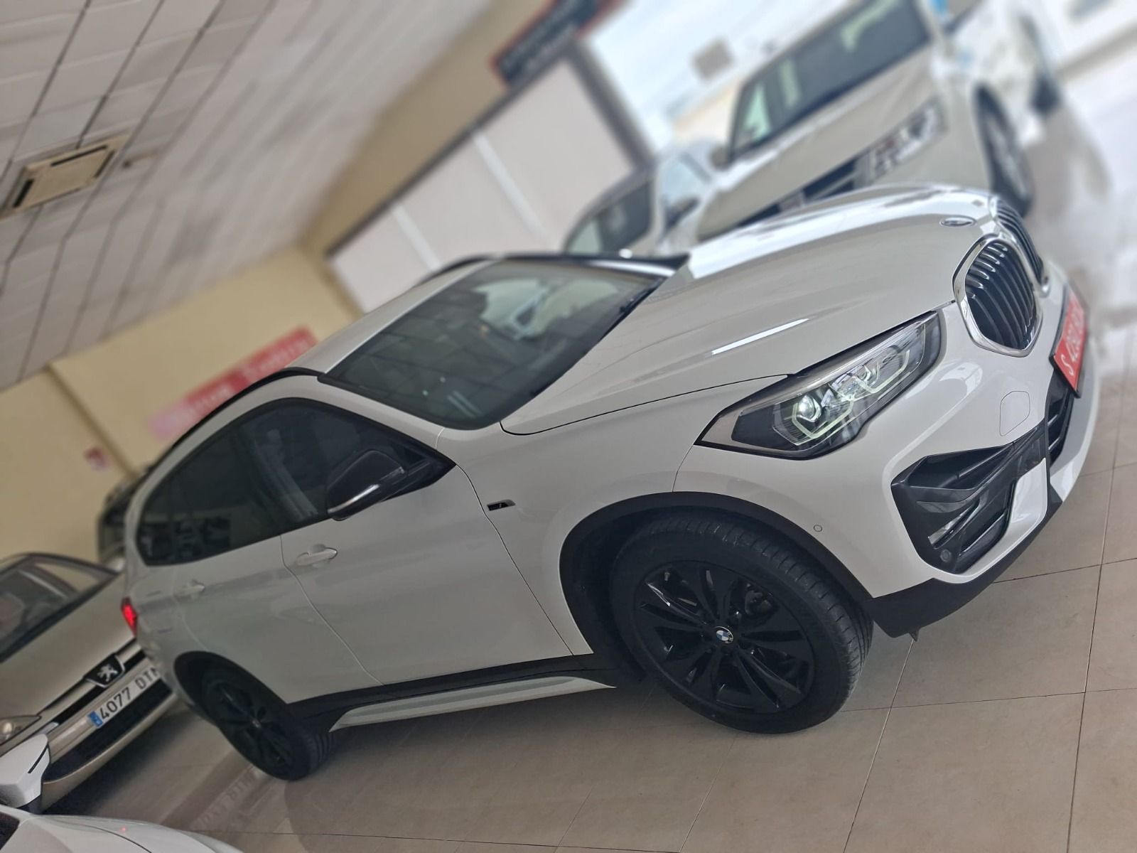 SUV BMW blanco estacionado en el interior sobre un suelo de hormigón, vista frontal en ángulo con ruedas negras.