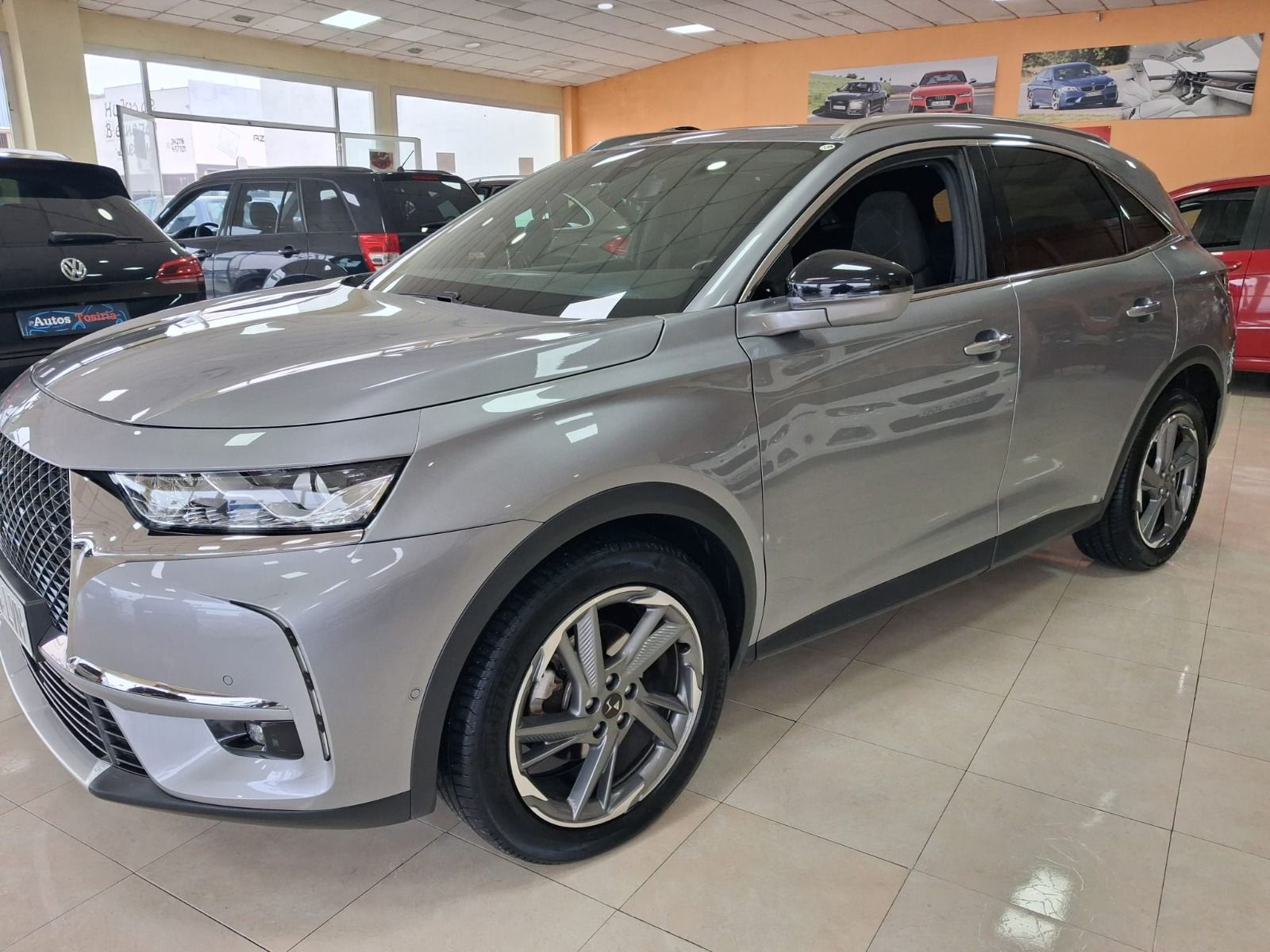 SUV plateado expuesto en una sala de exhibición, vista frontal izquierda, con otros vehículos al fondo.