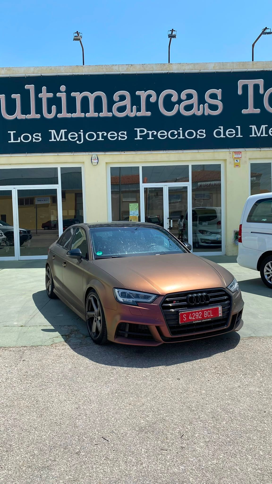 Un sedán deportivo color bronce estacionado frente a un concesionario de autos usados ​​con un letrero azul.