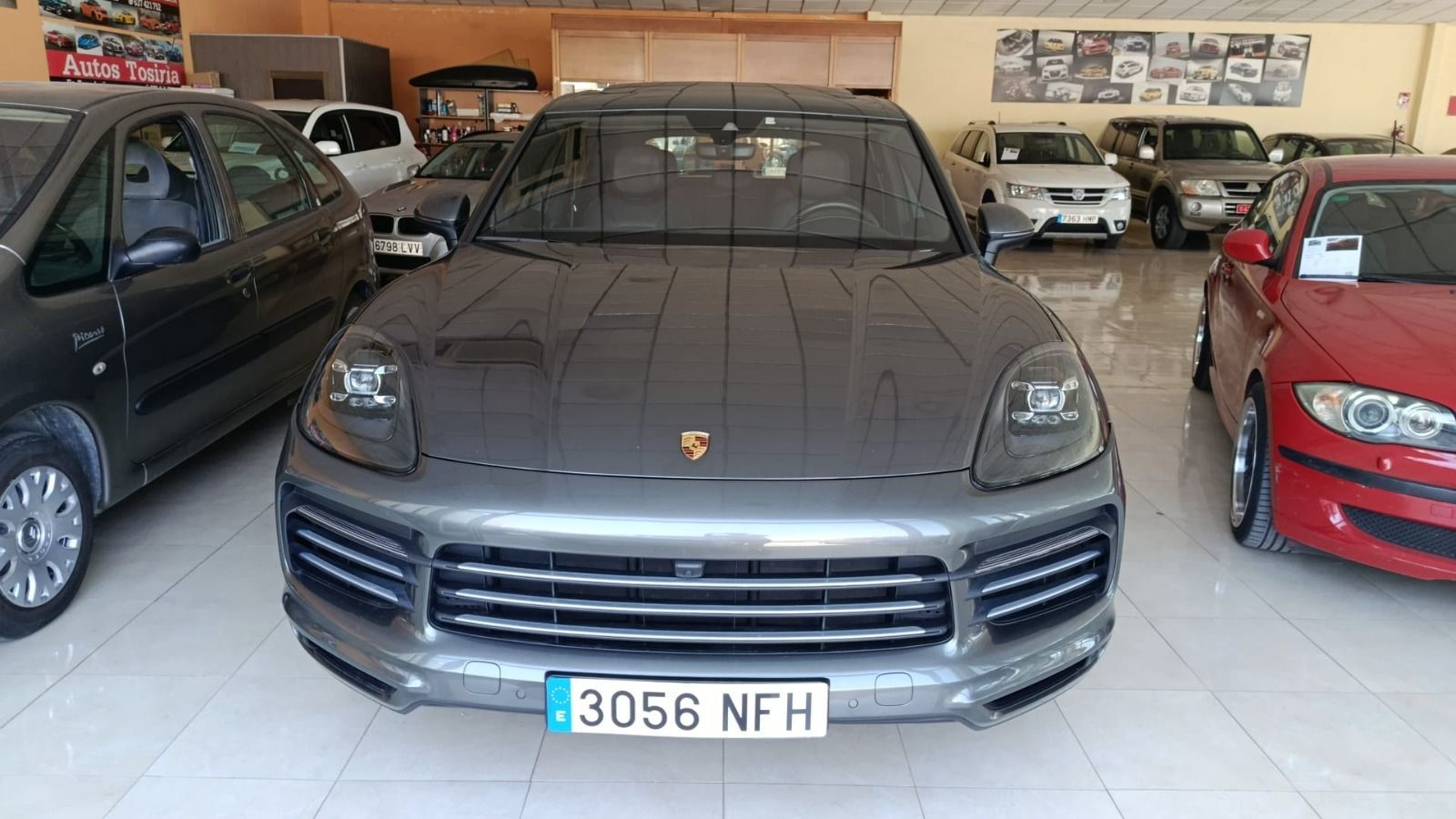 Vista frontal de un SUV Porsche gris estacionado en una sala de exposición, flanqueado por otros automóviles.