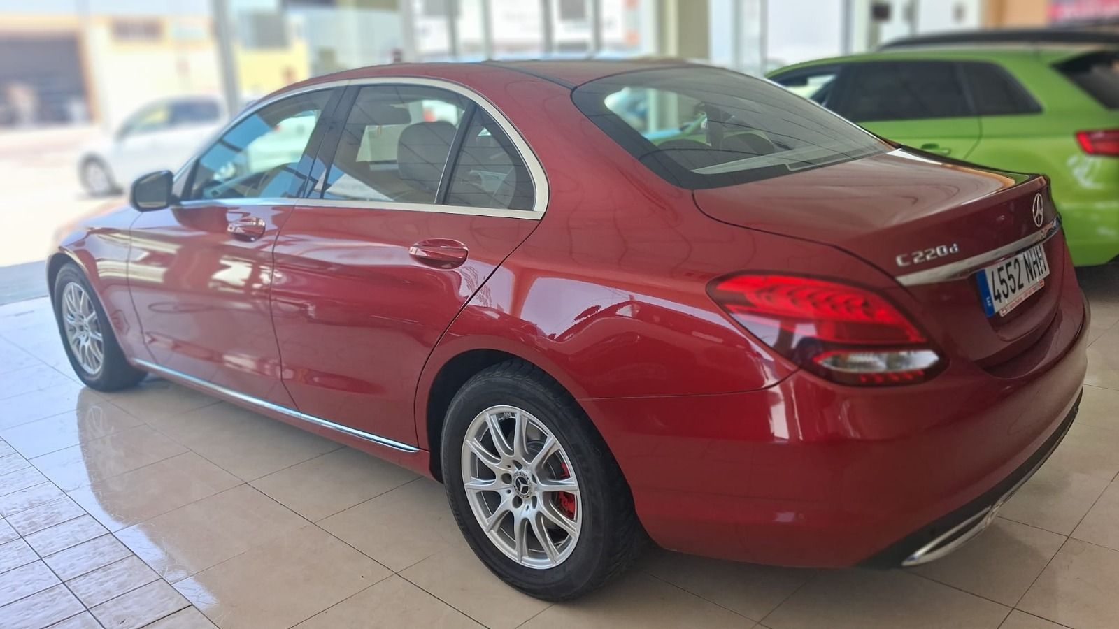 Sedán Mercedes-Benz rojo estacionado en el interior, vista trasera de tres cuartos, con un coche verde al lado.