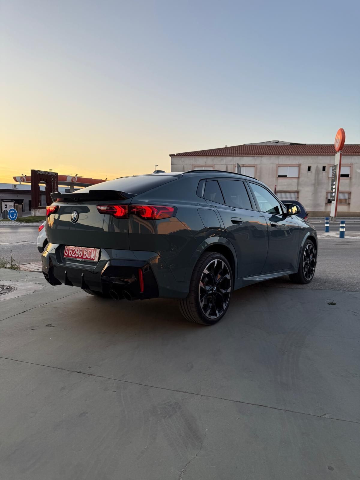 Sedán deportivo negro estacionado al atardecer en un lote industrial, vista trasera con luces traseras rojas visibles