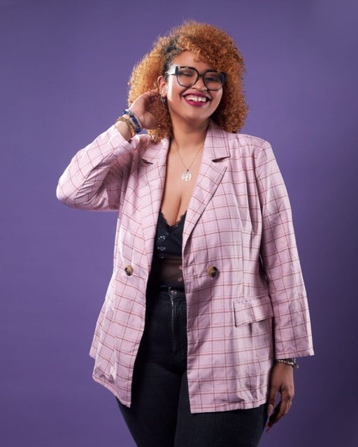 Una mujer que lleva gafas y una chaqueta rosa está parada frente a un fondo morado.