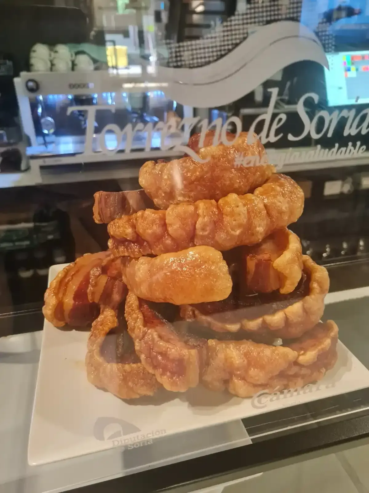 Montón de crujientes chicharrones dorados sobre un plato blanco, detrás de una vitrina de cristal en un bar.