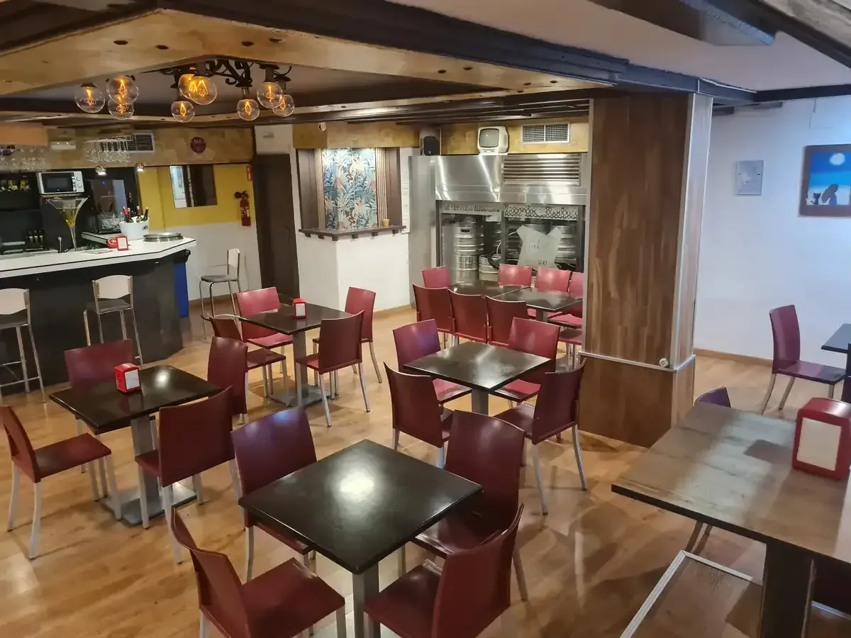 Interior de restaurante con mesas cuadradas, sillas rojas, barra y horno al fondo.