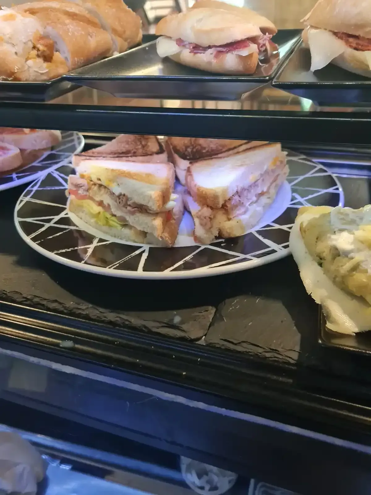 Sándwiches expuestos en una vitrina de cafetería. Uno de ellos está cortado por la mitad y colocado en un plato decorativo. Se ven los demás sándwiches.