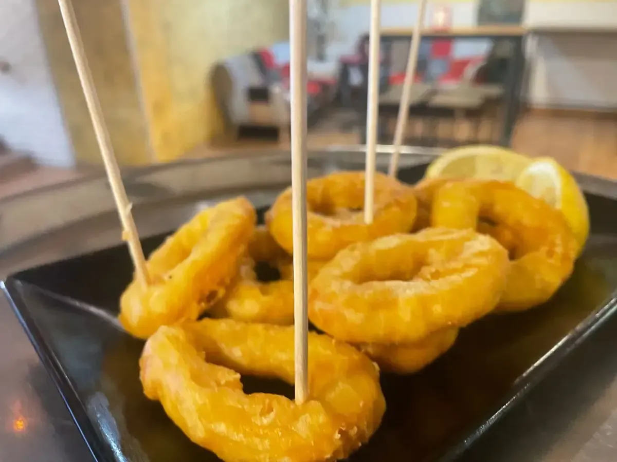 Aros de calamar fritos dorados sobre un plato negro, ensartados con palillos, una rodaja de limón.