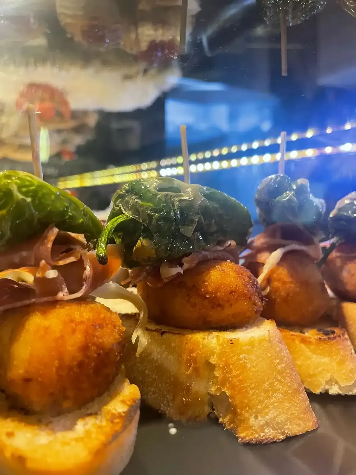 Primer plano de tapas: bolas fritas sobre rebanadas de pan, cubiertas con verduras, carne y brochetas.