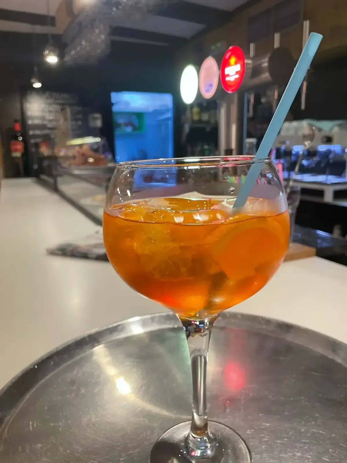 Cóctel de naranja servido en copa con pie y pajita azul sobre una bandeja de plata en un bar.