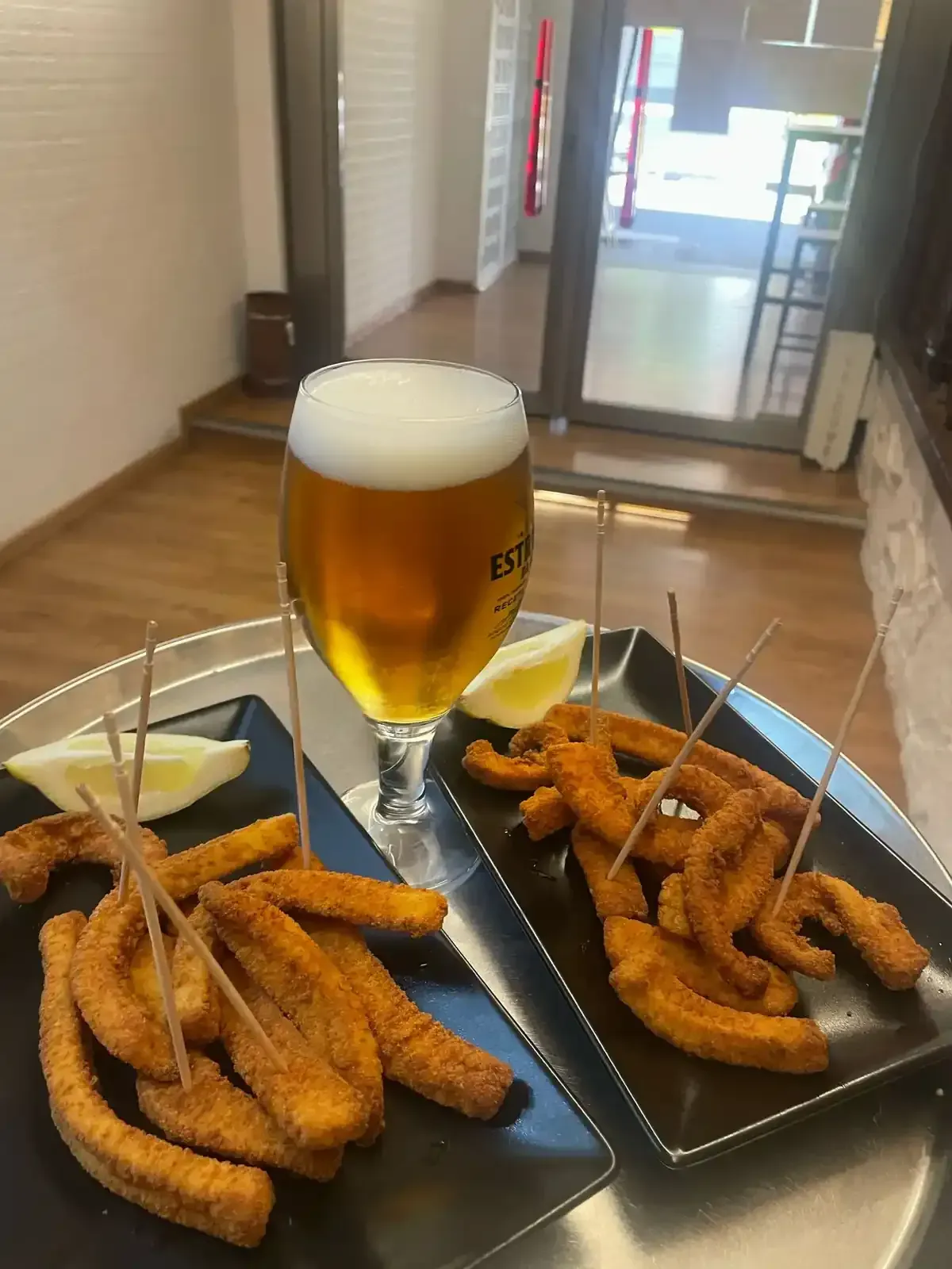Un vaso de cerveza con espuma, comida frita en dos platos negros y rodajas de limón sobre una mesa de metal.