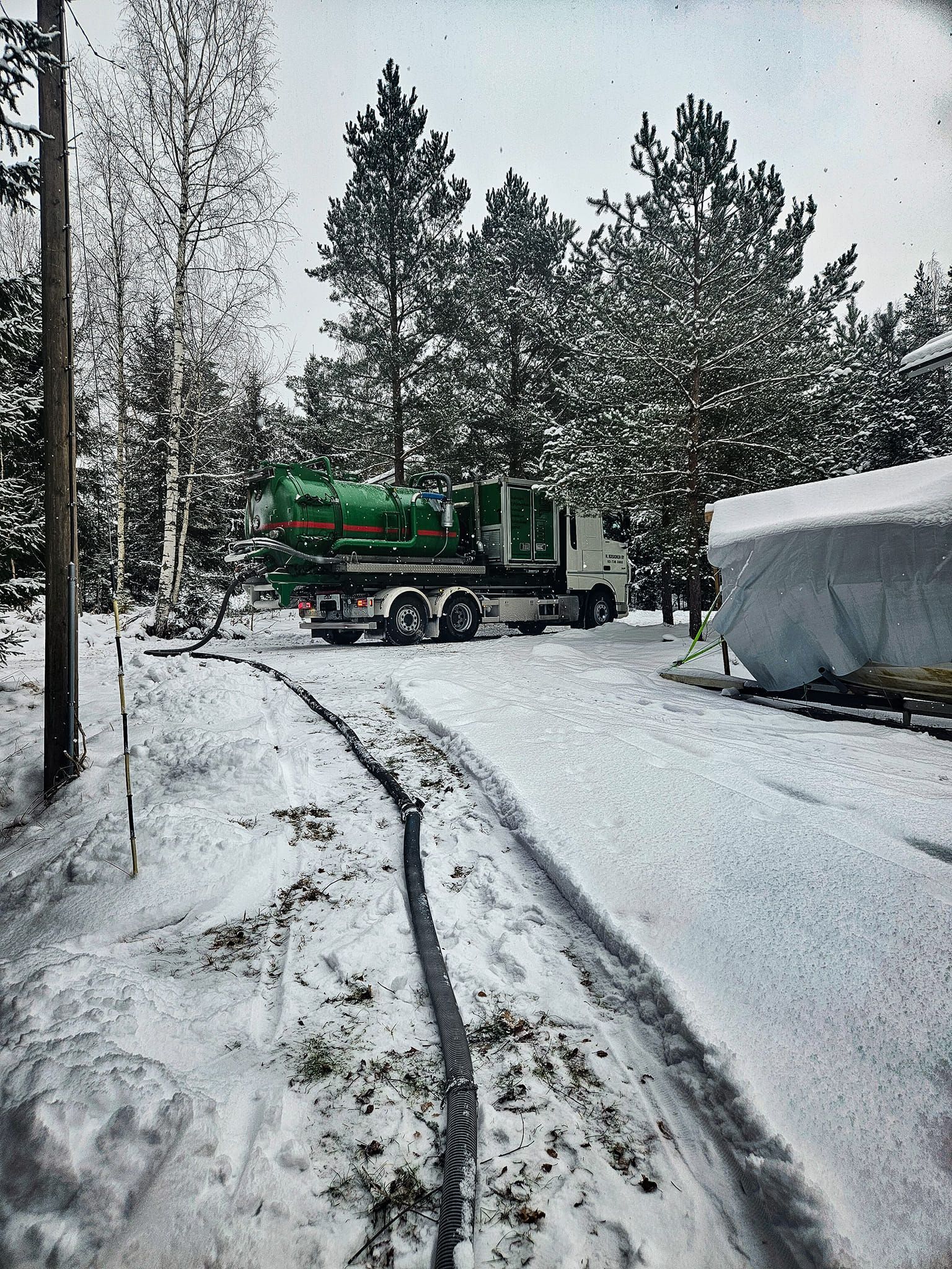 Luminen maisema: Vihreä säiliöauto pysäköitynä talviselle ajotielle, letku johtaa peitettyyn säiliöön.