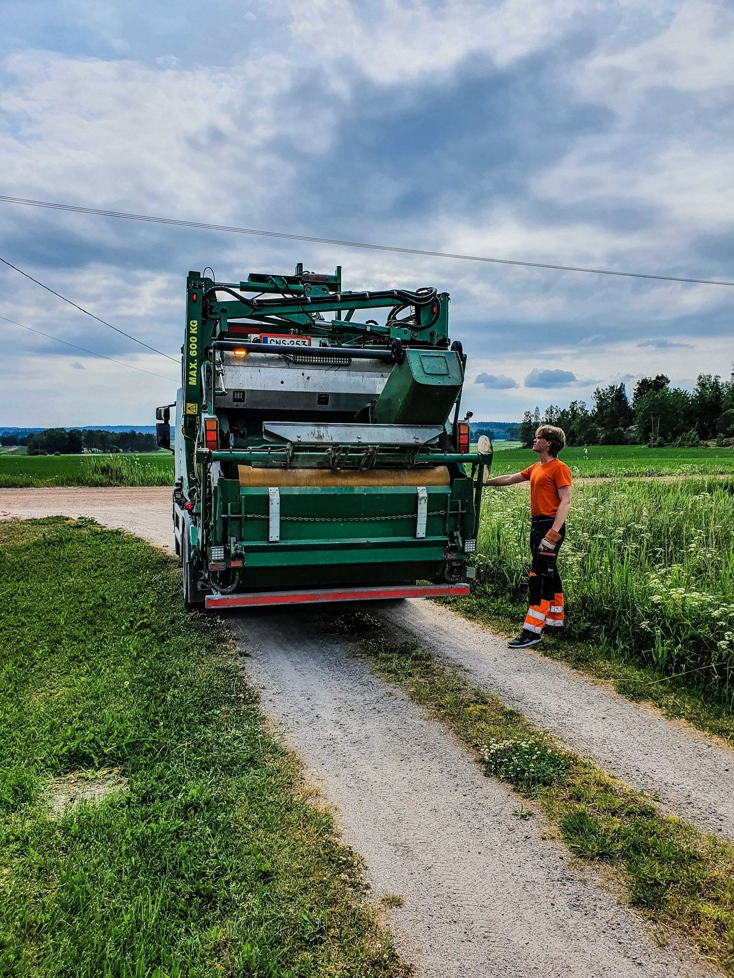 Roska-auto soratietä pitkin, ja sen vieressä pellolla seisoo oranssiin ja mustaan ​​pukeutunut henkilö. Pilvinen taivas.