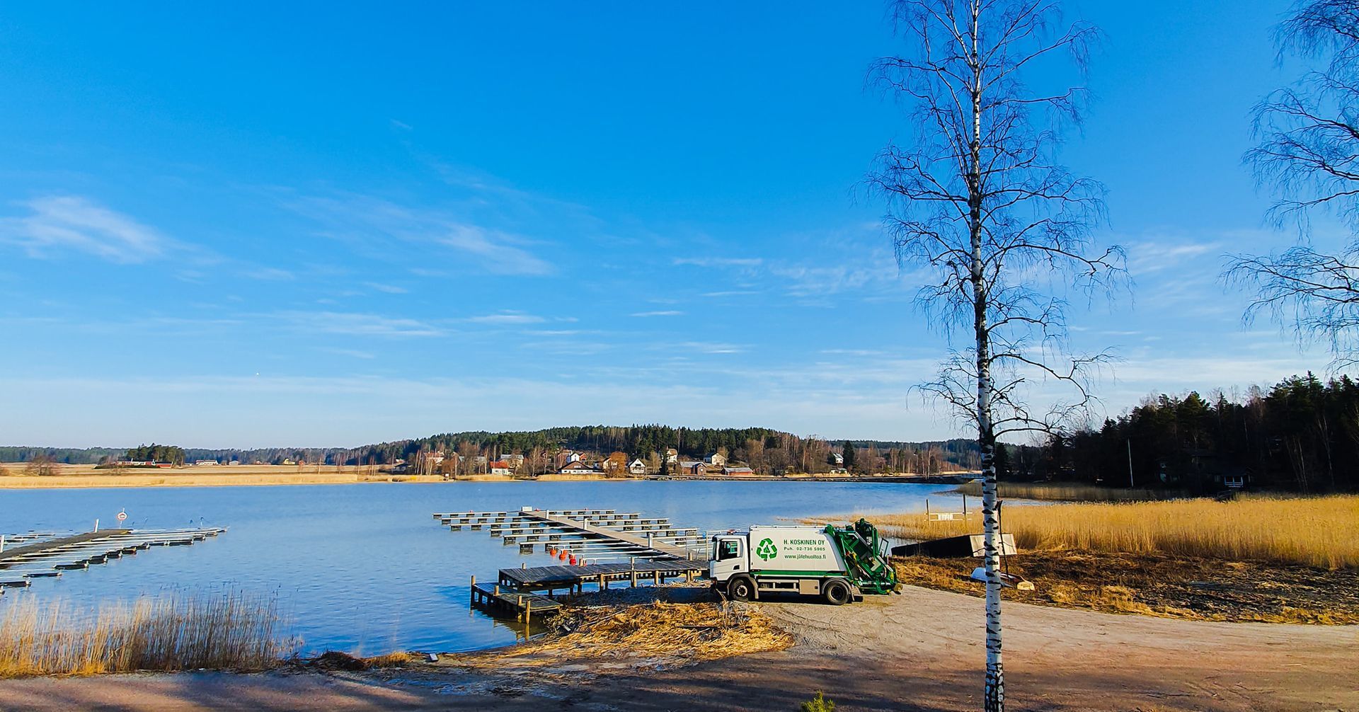 Järvenrantanäkymä laitureilla, puilla ja vihreällä jäteautolla kirkkaan sinisen taivaan alla.
