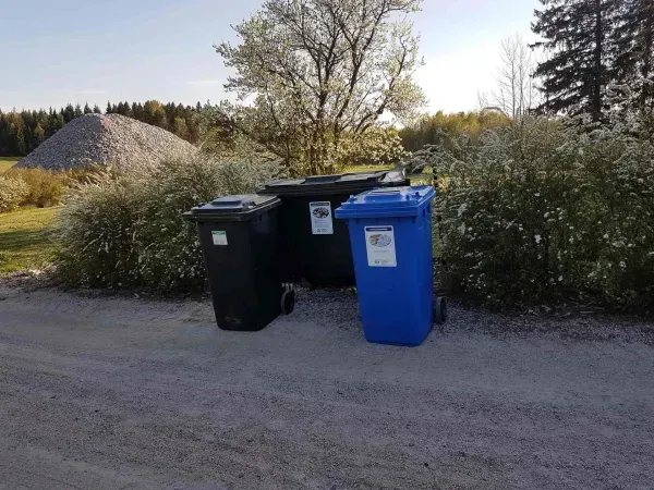 Kolme kierrätysastiaa, mustaa ja sinistä, sorapolulla, taustalla lehtiä ja puuta.