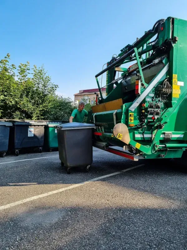 Roska-auto keräämässä tummaa roskakoria. Vihreäpukuinen työntekijä seisoo lähellä sinisen taivaan alla.