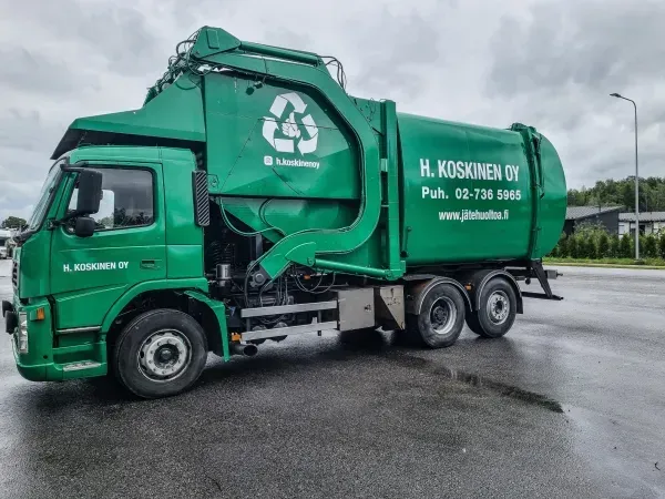 Vihreä jäteauto kierrätyssymbolilla pysäköitynä märälle asfaltille.