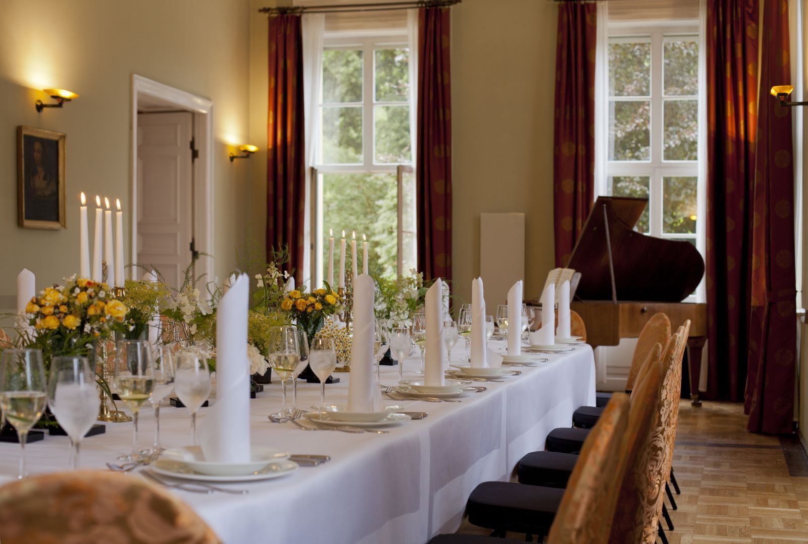 Ein langer Esstisch ist für eine Mahlzeit gedeckt, mit Blumengestecken, Kerzen und einem Klavier im Hintergrund.