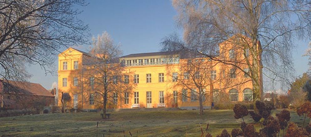 Gelbes Gebäude mit vielen Fenstern, Bäumen und Gras unter blauem Himmel.