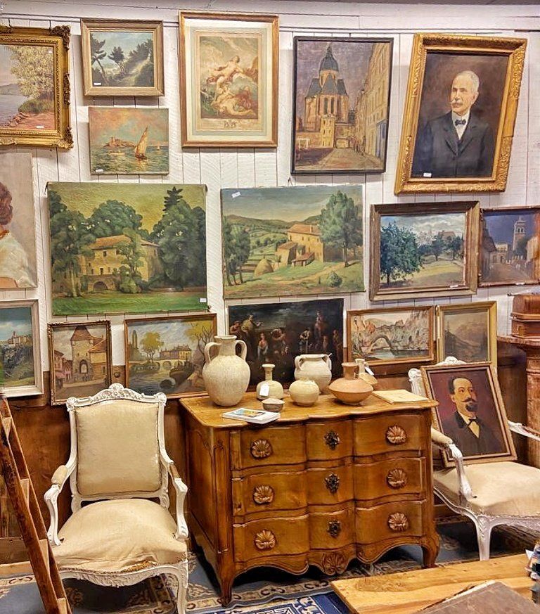Mur chez un antiquaire recouvert de tableaux, avec deux chaises et un meuble à tiroir devant
