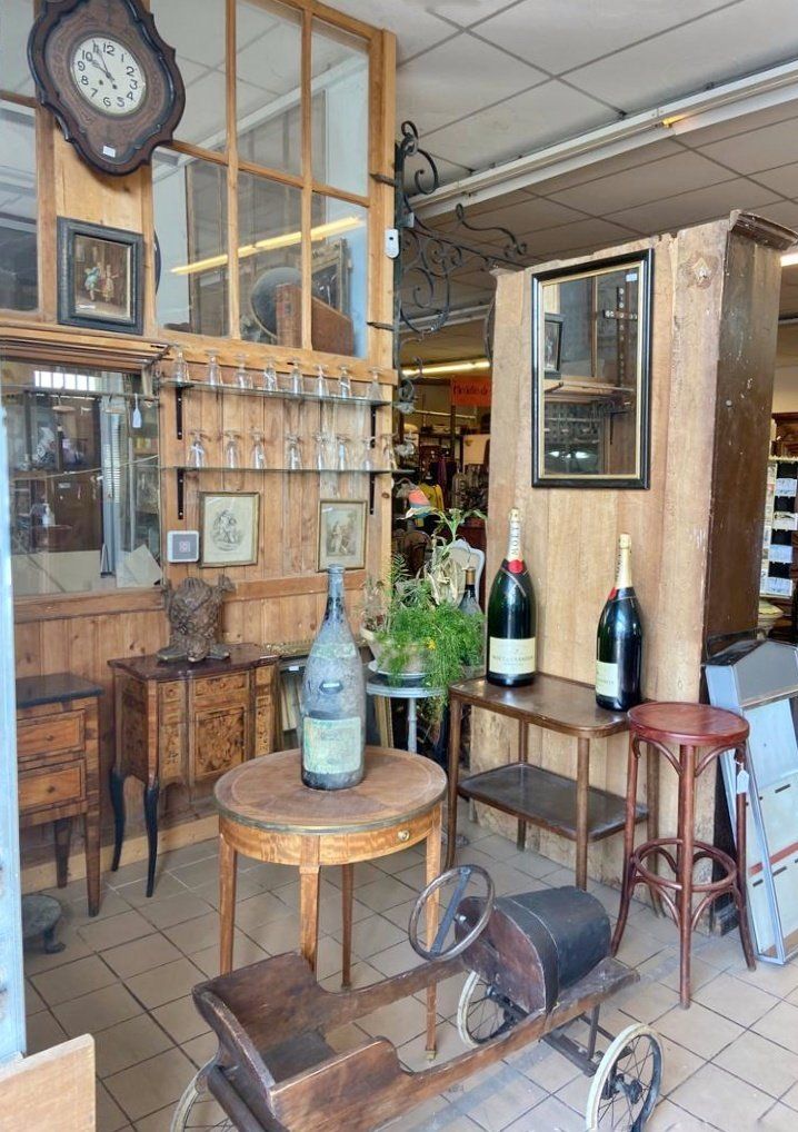 Trois bouteilles de vin sur deux tables différentes, avec une auto d'enfant en bois devant, des verres rangés derrière