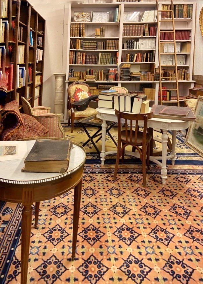 Coin Librairie d'un antiquaire avec des étagères de livres à gauche et au centre, et deux tables et des sièges au centre