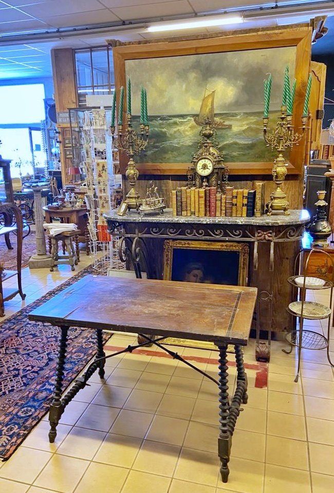 Table en bois sans rien dessus devant une desserte portant des livres, une horloge, deux candélabres, devant un grand tableau