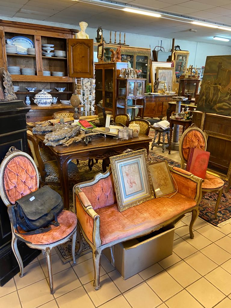 Espace d'un antiquaire avec deux chaises et un canapé entre, sur lequel reposent deux tableaux, devant une table en bois