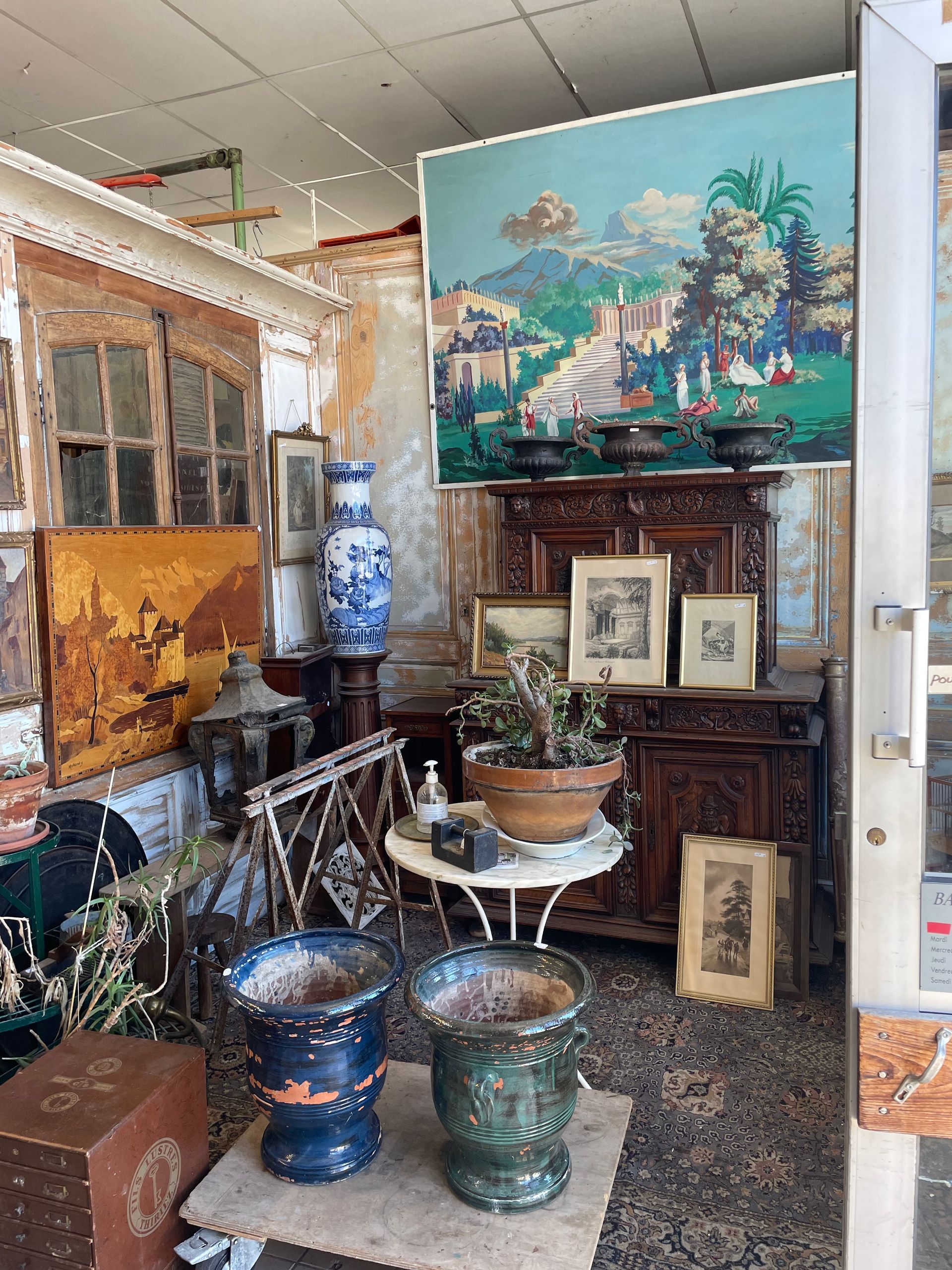 Chez un antiquaire une table de jardin en fer devant un meuble en bois, et à gauche un tableau derrière une lanterne en fer