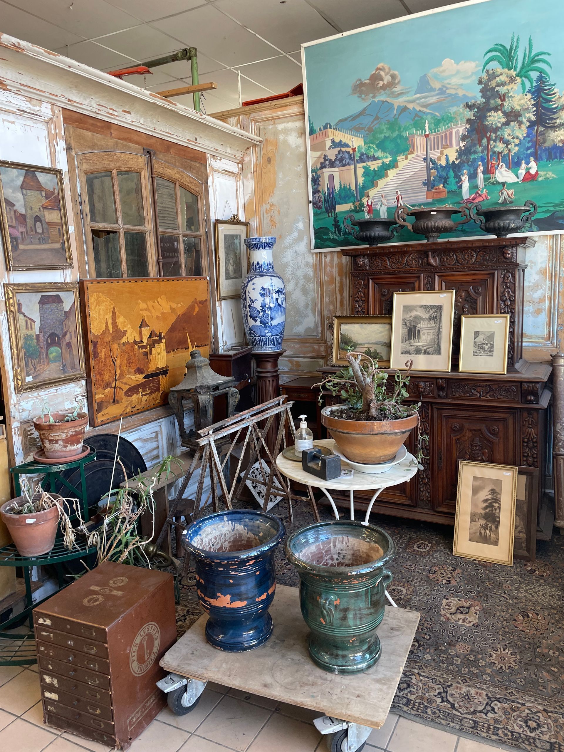 Espace chez un antiquaire avec des tableaux aux murs, des pots à fleurs au premier plan, et une table en fer au centre