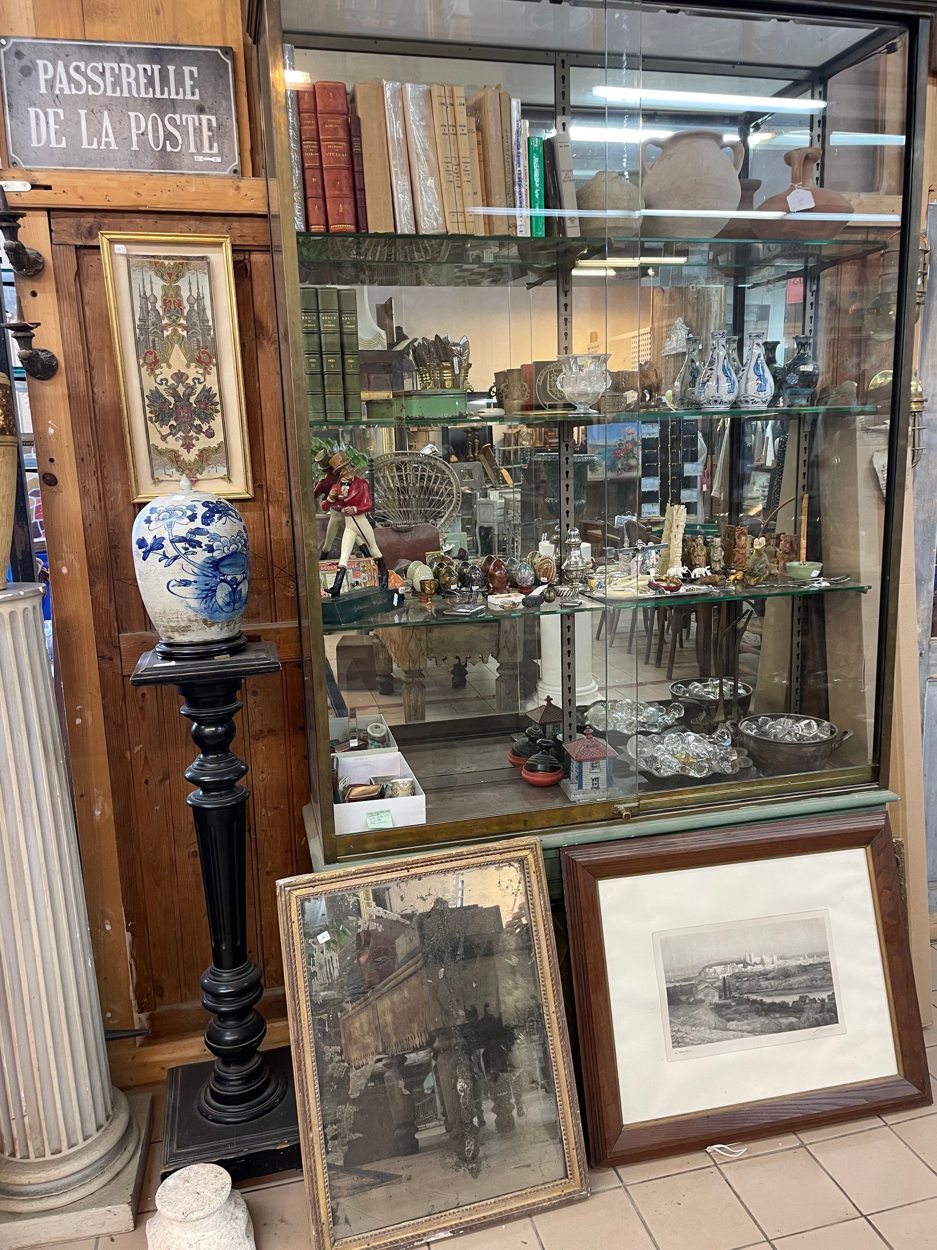 Vitrine en verre contenant des objets décoratifs avec des tableaux en bas, et à gauche au dessus d'un pot