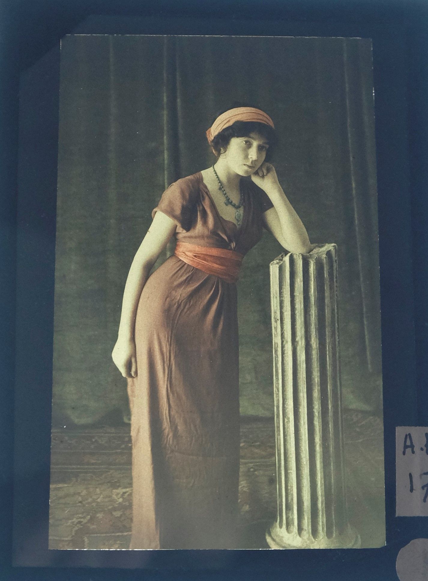 Photographie montrant une femme en robe marron orange debout accoudée à une colonne grecque la tête sur le poing