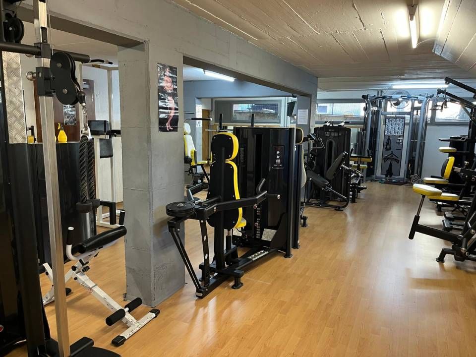 Vue intérieure d'une salle de sport avec divers appareils de musculation noirs et jaunes disposés sur un parquet clair.