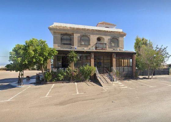 Edificio de dos plantas con porche de madera y aparcamiento, bajo un cielo azul claro.