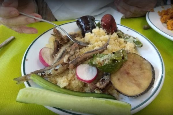 Plato de cuscús, sardinas, verduras y una mano sujetando un tenedor.