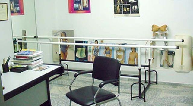 Sala de fisioterapia con barras paralelas, una silla y aparatos de ejercicio frente a una pared de espejos.