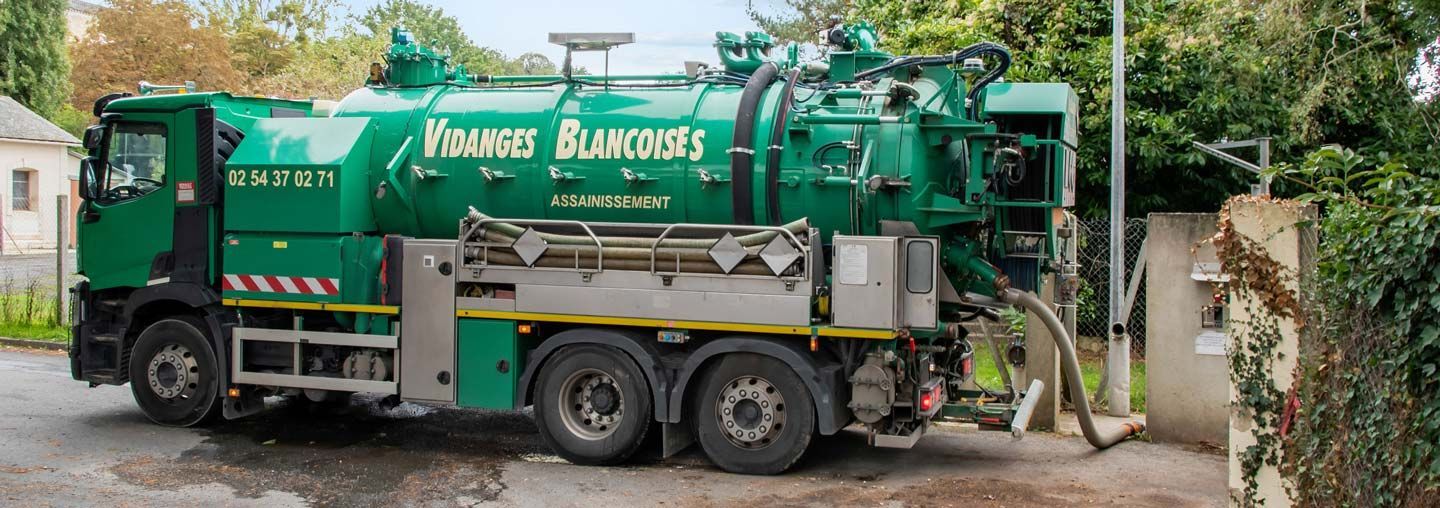 Un camion aspirateur vert est garé près d’une haie, avec un tuyau déployé en direction du mur d’une maison.