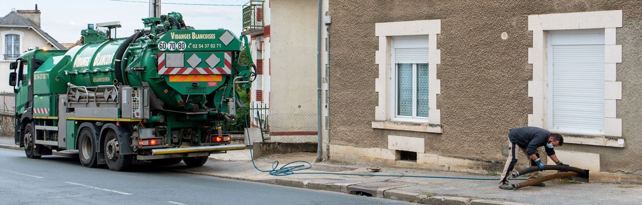 Un camion-benne vert est garé dans une rue étroite tandis qu’un employé balaie le trottoir près d’un bâtiment en briques.