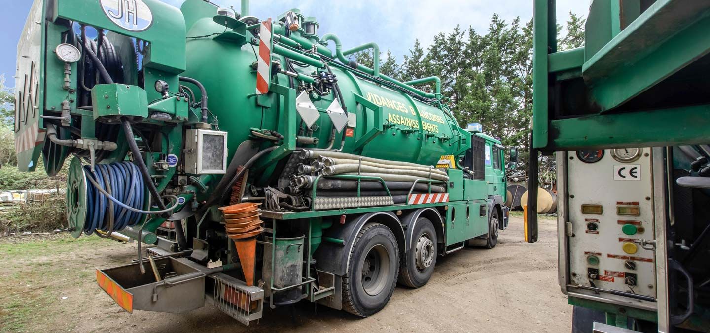 Camion aspirateur industriel vert stationné à l’extérieur sur un terrain de gravier, avec des tuyaux et du matériel fixés au véhicule