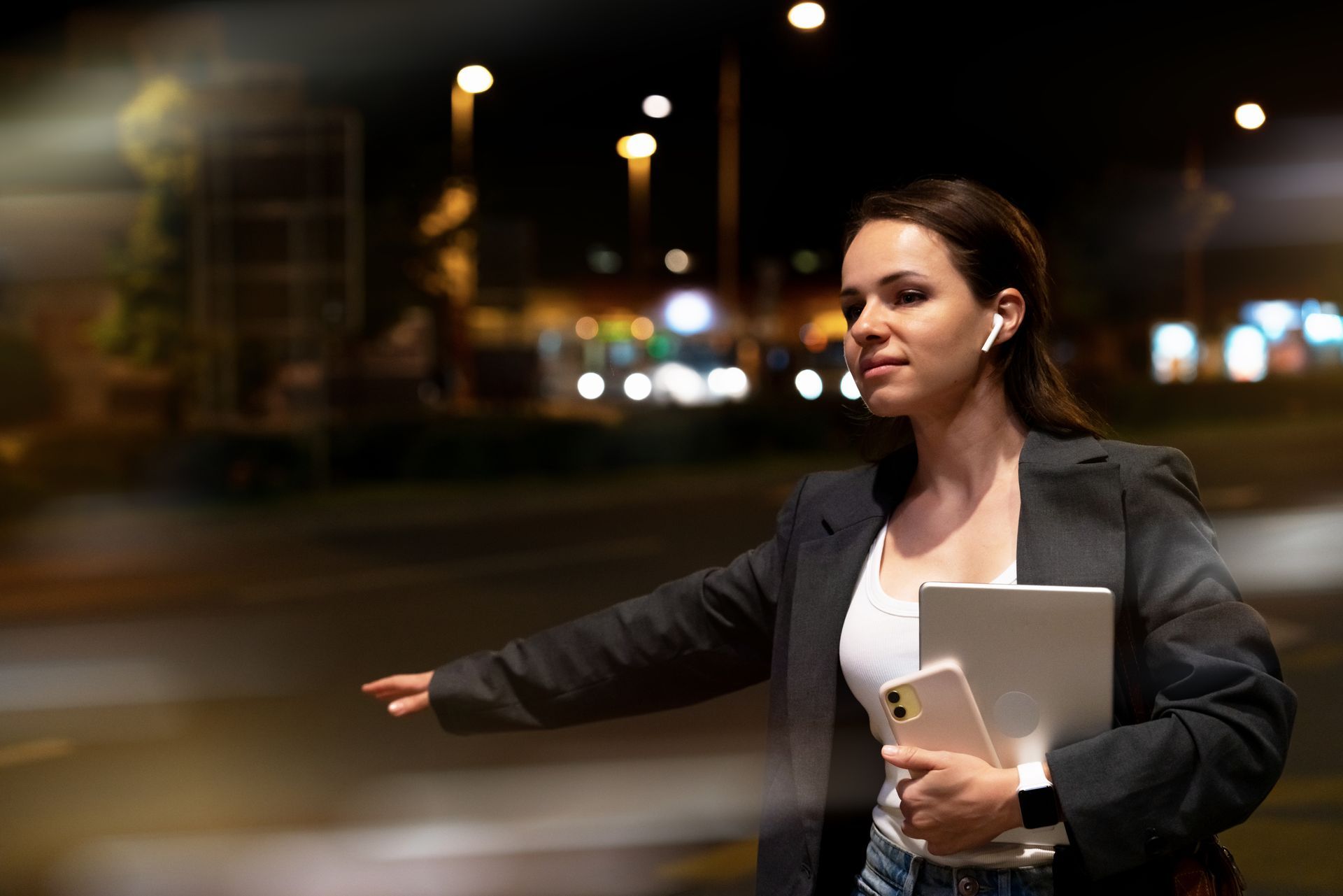 Une femme en blazer tient son téléphone et son ordinateur portable, hélant un taxi de nuit.