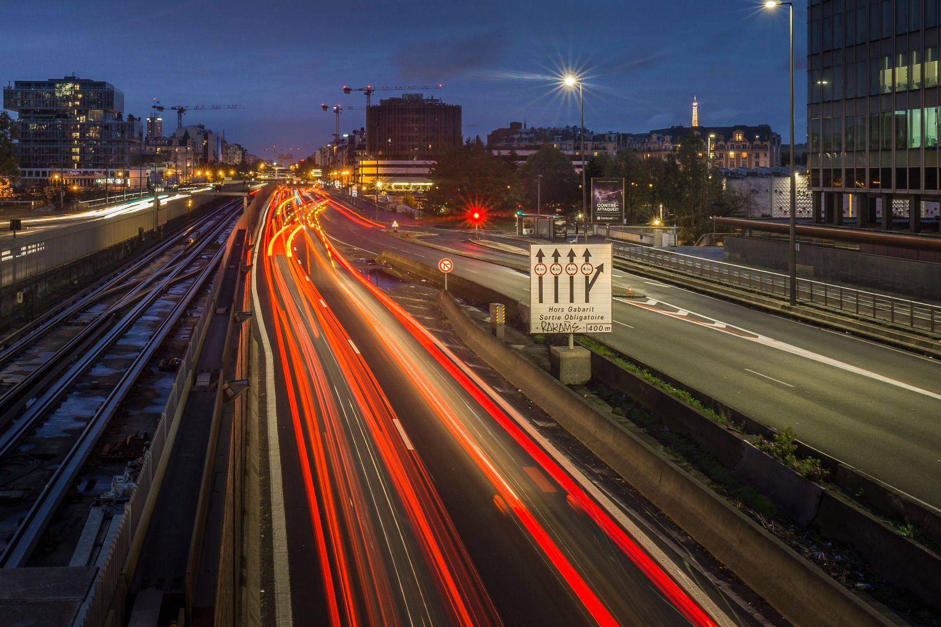 Lumières de la ville au crépuscule ; longue exposition de traînées de circulation sur l’autoroute. Voie ferrée parallèle. Bâtiments en arrière-plan.