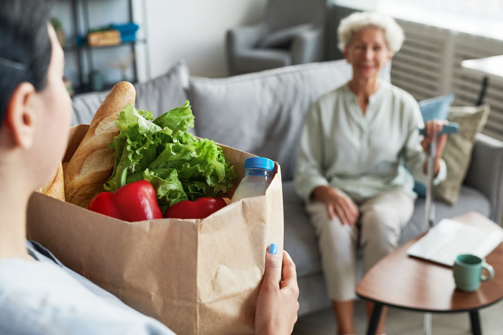 Une femme livre des courses à une personne âgée assise sur un canapé.