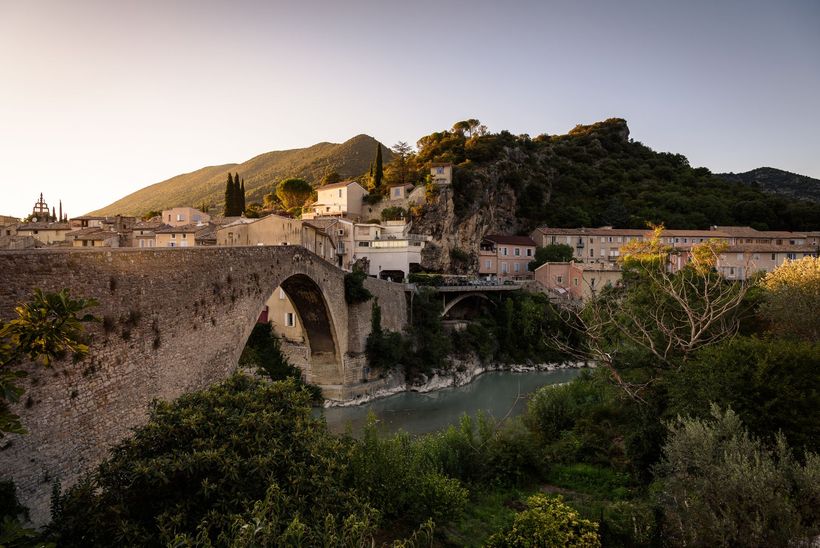 Un vieux pont de pierre et un village niché près d'une rivière, avec des bâtiments et des montagnes en arrière-plan.