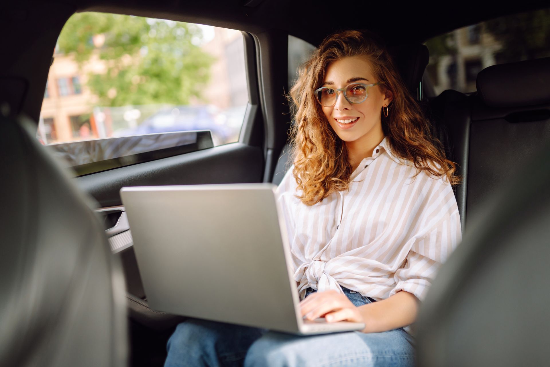 Une femme portant des lunettes travaille sur son ordinateur portable sur la banquette arrière d'une voiture.
