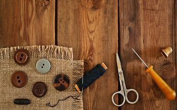 Suministros de costura sobre una mesa de madera: botones, hilo, tijeras, aguja, dedal y arpillera.
