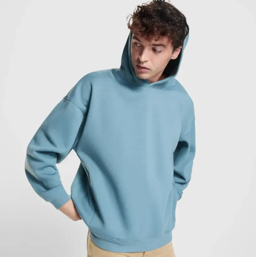 Hombre con sudadera azul y pantalones color canela, mirando hacia un lado, con las manos en los bolsillos.