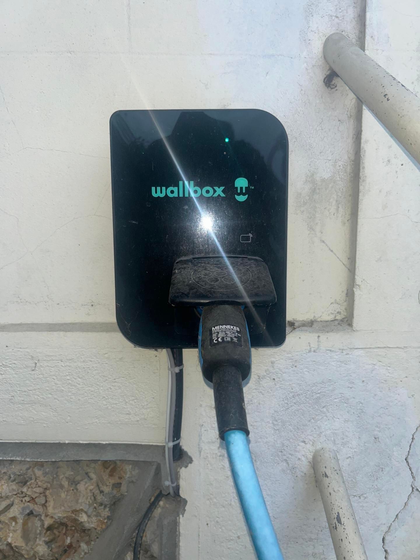 Une borne de recharge murale noire pour véhicules électriques, fixée sur un mur blanc, avec un câble de recharge bleu branché dessus.